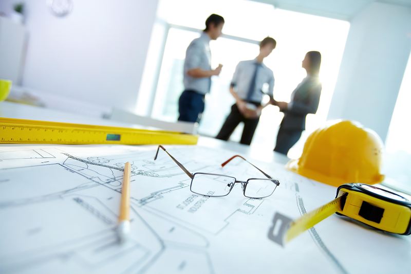 Construction drawings spread across a table with a tape measure, pencil, safety glasses and a hard hat, three colleagues talking in the background