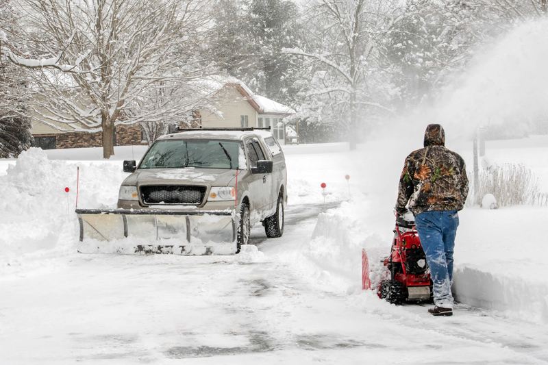Snow blower clearing a driveway walkway beside plowed banks, with a plow truck on a snow-covered residential street