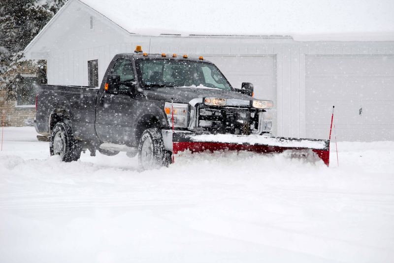 Pickup truck with a red plow blade pushing snow past orange marker stakes in front of a commercial building