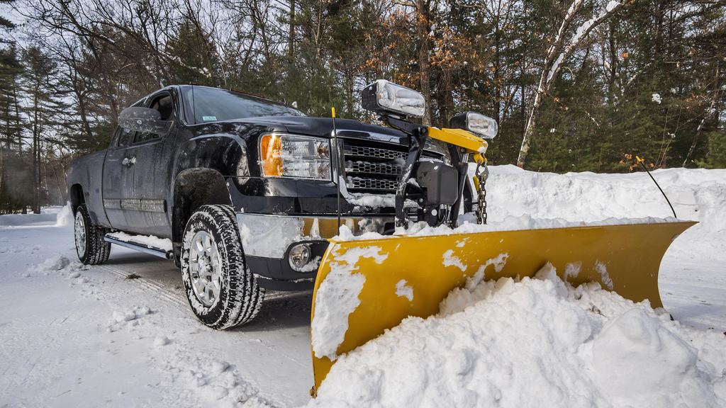 Pickup truck fitted with a yellow snow plow blade pushing snow beside a treeline